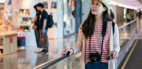 Woman in a striped shirt and straw hat pulling a suitcase through an airport terminal with a camera around her neck.