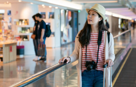 Woman in a striped shirt and straw hat pulling a suitcase through an airport terminal with a camera around her neck.