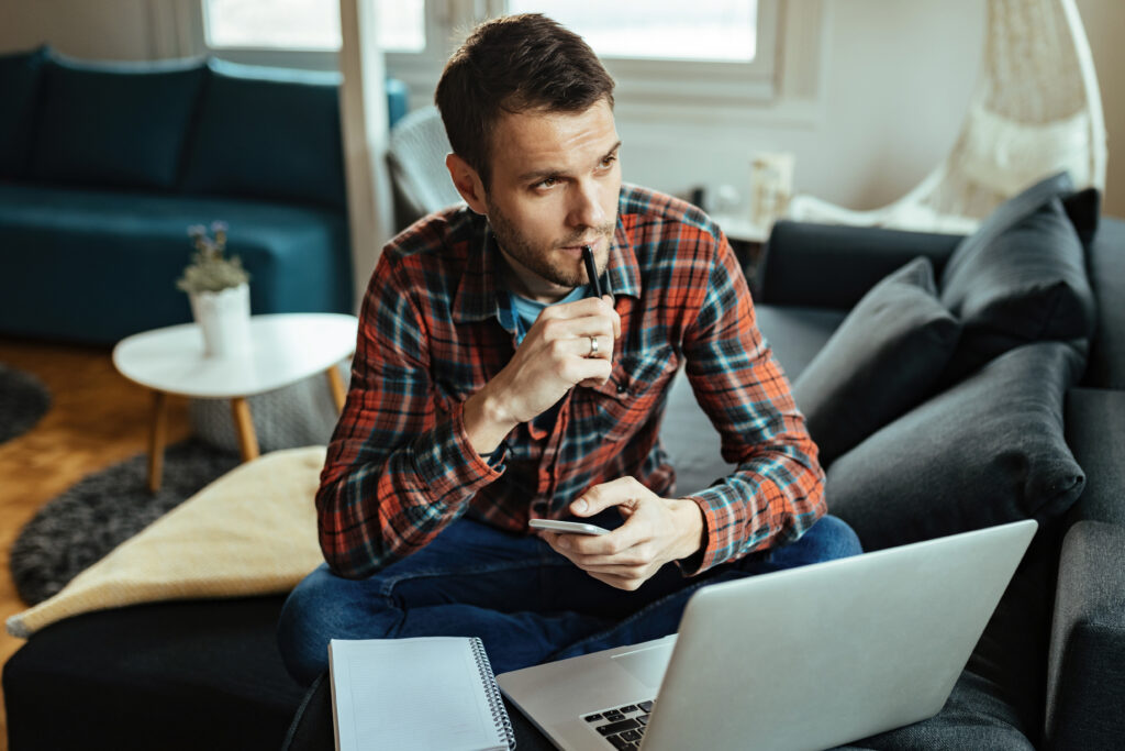 Man sitting on a couch with a laptop and notebook, holding a phone and thinking with a pen to his lips.