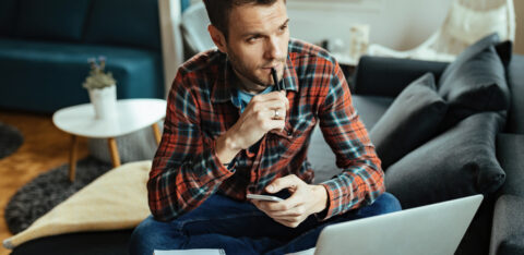 Man sitting on a couch with a laptop and notebook, holding a phone and thinking with a pen to his lips.