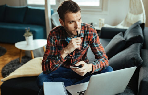 Man sitting on a couch with a laptop and notebook, holding a phone and thinking with a pen to his lips.
