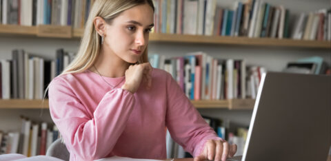 Focused young woman in a pink sweater studying at a desk with a laptop and open books.