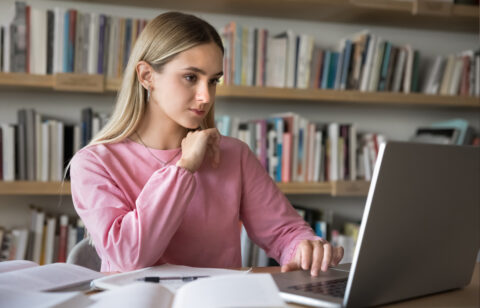 Focused young woman in a pink sweater studying at a desk with a laptop and open books.