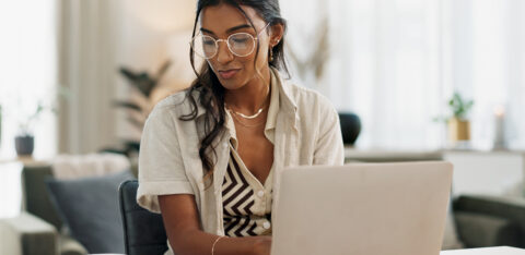 Woman working at a laptop while writing notes in a notebook at her desk.
