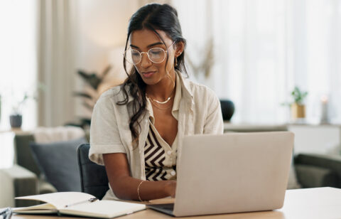 Woman working at a laptop while writing notes in a notebook at her desk.