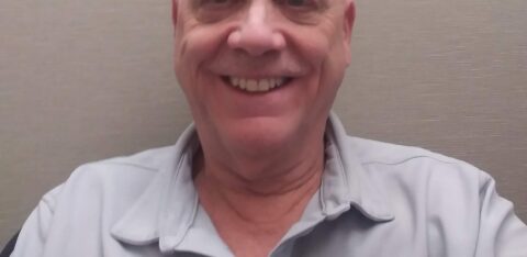 Smiling older man wearing a light gray collared shirt, seated in front of a neutral background.