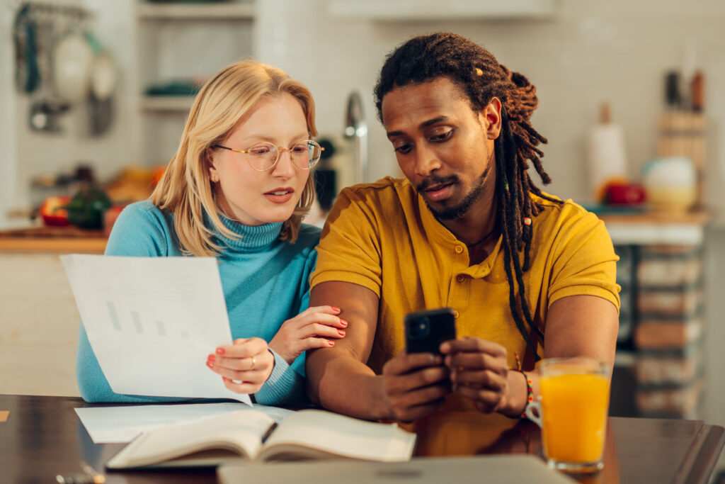 Couple sitting at a kitchen table reviewing financial documents together, with a phone and open notebook in front of them.