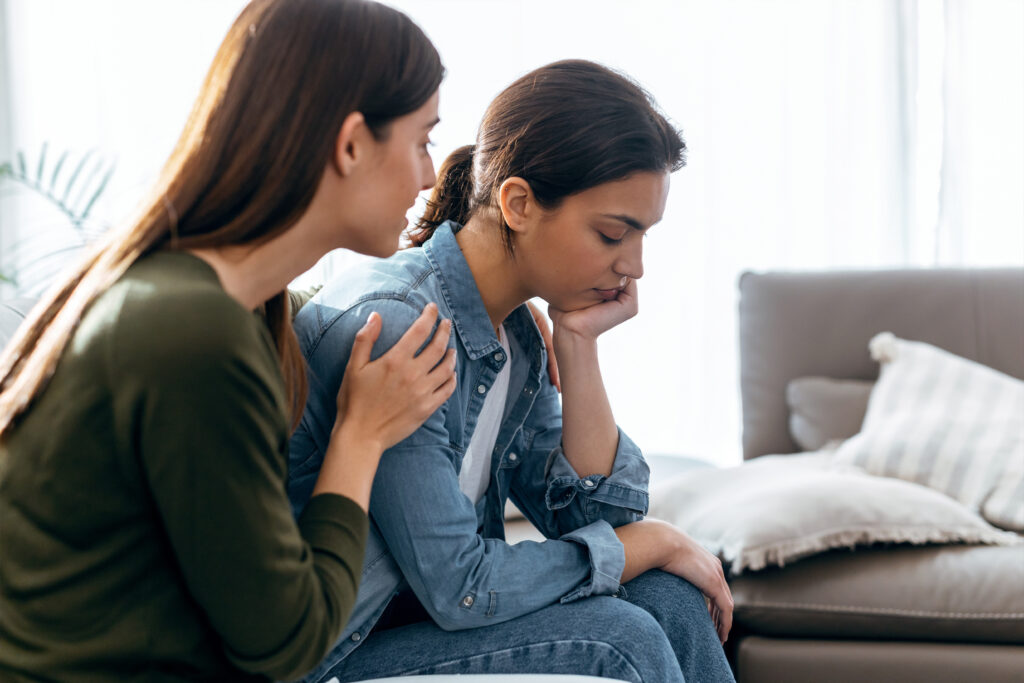 A woman sits with her head resting on her hand, looking upset, while another woman gently comforts her by placing a hand on her shoulder.