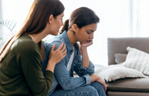 A woman sits with her head resting on her hand, looking upset, while another woman gently comforts her by placing a hand on her shoulder.