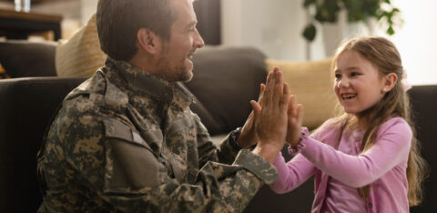 A smiling man in military uniform playing a clapping game with his young daughter in a bright living room.