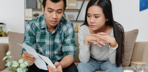 A couple sitting on the couch reviewing bills and financial documents with serious expressions.