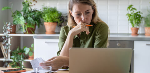 Woman sitting at a kitchen table, reviewing bills and using a laptop with a thoughtful expression.