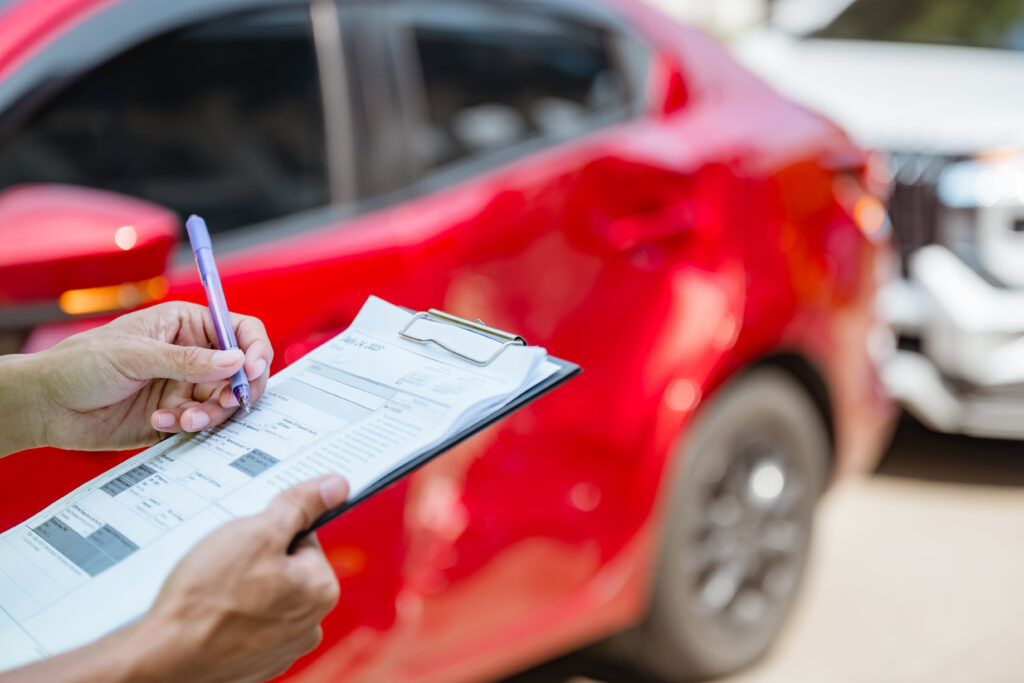 Person filling out an insurance claim form on a clipboard near a red car after an accident