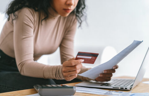 Woman reviewing a bill while holding a credit card, with a calculator and laptop on a desk.