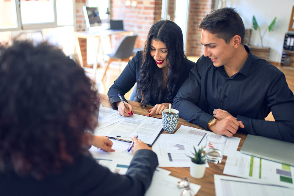 A smiling couple meets with a professional, reviewing and signing financial documents at a desk with charts and paperwork.