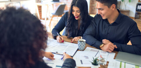A smiling couple meets with a professional, reviewing and signing financial documents at a desk with charts and paperwork.