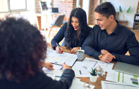 A smiling couple meets with a professional, reviewing and signing financial documents at a desk with charts and paperwork.