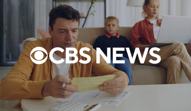 A parent reviews bills at home with a partner and child in the background, with the CBS News logo overlayed across the image