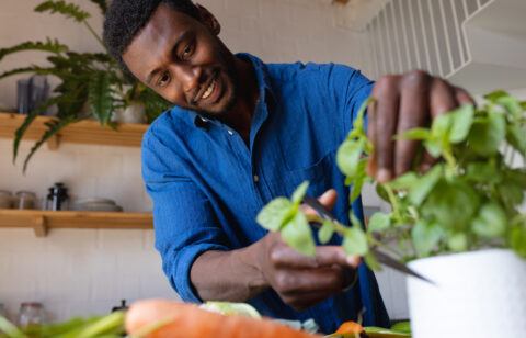Smiling man in a blue shirt trimming fresh herbs in a kitchen with vegetables on the counter.