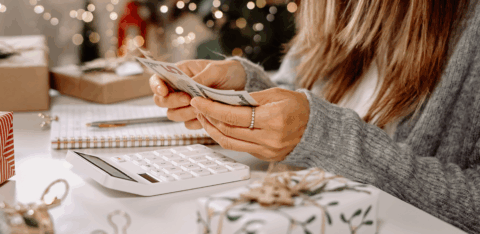 Woman counting cash while planning a holiday budget with gifts and decorations on the table.