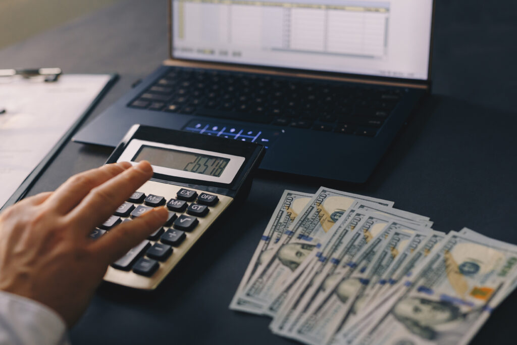 Hand pressing calculator buttons next to cash and a computer displaying a financial spreadsheet.