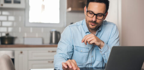 A man using a calculator while working on a laptop at his kitchen table.