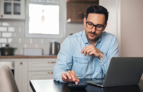 A man using a calculator while working on a laptop at his kitchen table.