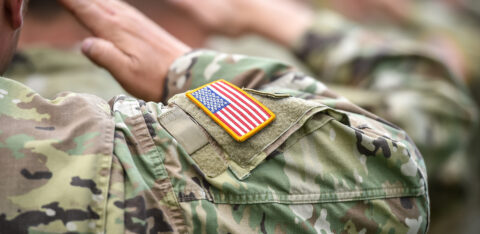 U.S. service members in camouflage uniforms saluting, with an American flag patch visible on one sleeve.