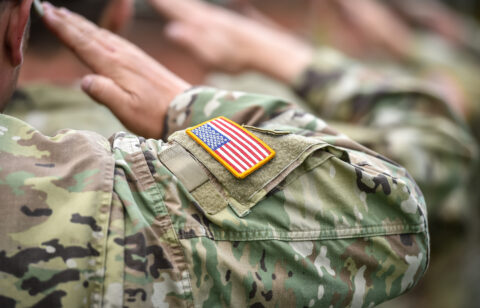 U.S. service members in camouflage uniforms saluting, with an American flag patch visible on one sleeve.