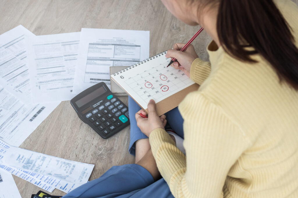 Person sitting on the floor with bills, a calculator, and a calendar, marking due dates for payments.