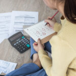 Person sitting on the floor with bills, a calculator, and a calendar, marking due dates for payments.
