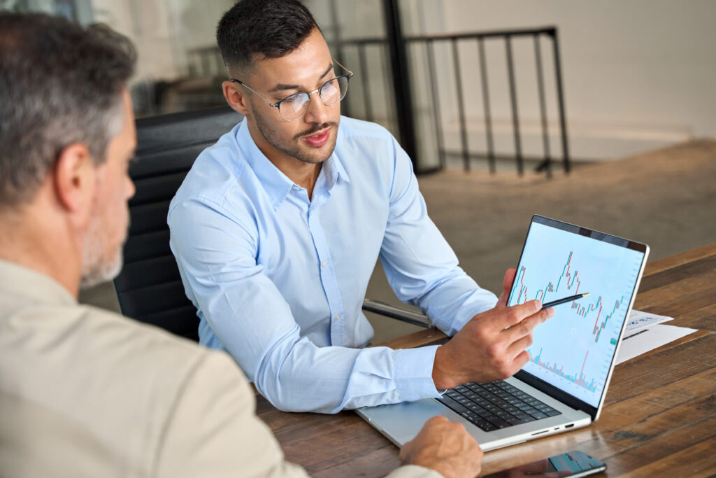 Man in glasses pointing at a financial graph on a laptop while discussing data with a colleague