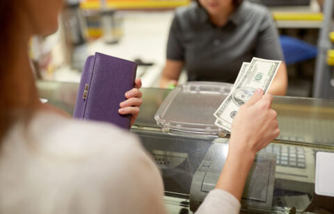A person hands cash to a cashier across a counter while holding a purple wallet.