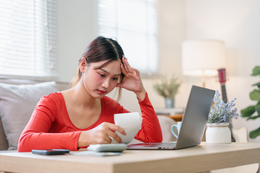 A young woman sits at a table with her laptop and calculator, looking worried as she reviews a bill at home.