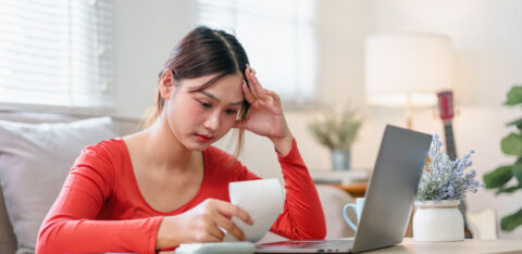 A young woman sits at a table with her laptop and calculator, looking worried as she reviews a bill at home.