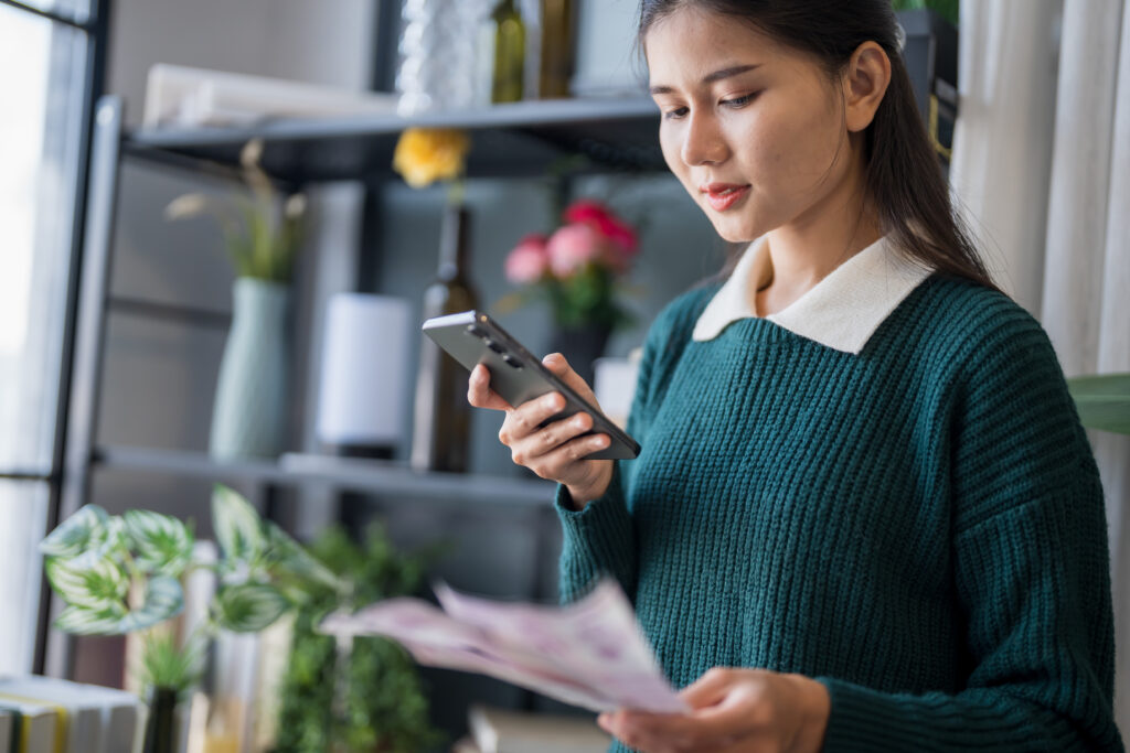 Young woman using her phone to review bills or financial documents at home.