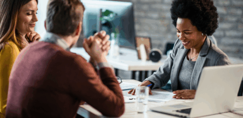 A smiling business professional meets with two clients at a modern office table, reviewing paperwork together.