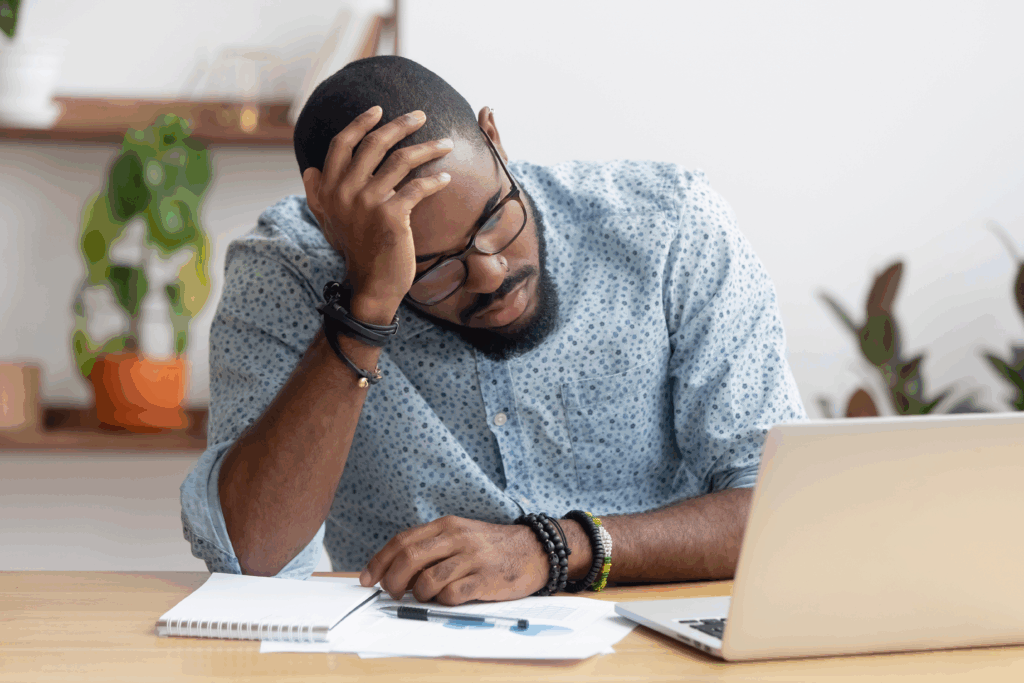 Stressed man sitting at a desk with a laptop, holding his head while reviewing financial papers.