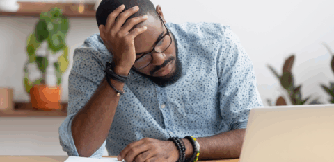 Stressed man sitting at a desk with a laptop, holding his head while reviewing financial papers.