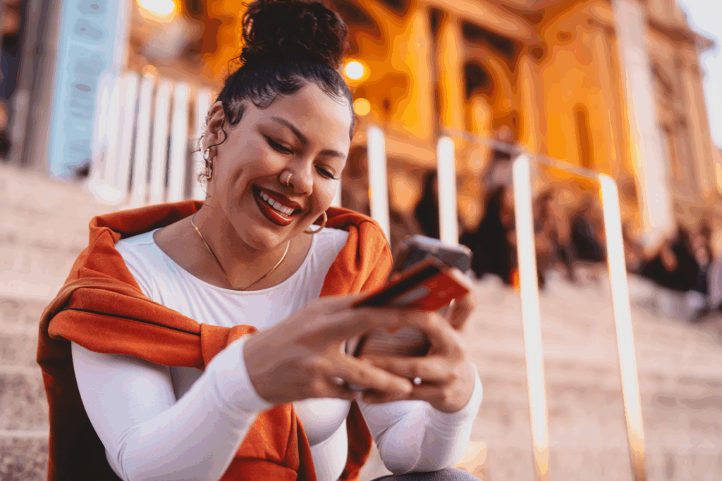 A smiling woman sits on outdoor steps in the evening, holding a phone and credit card while looking at her screen.