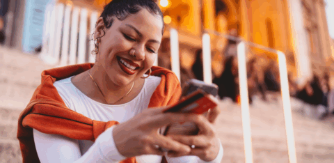 A smiling woman sits on outdoor steps in the evening, holding a phone and credit card while looking at her screen.