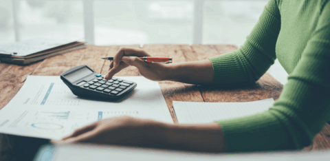 A person wearing a green sweater uses a calculator and reviews financial charts at a wooden desk.