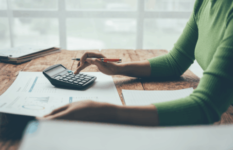 A person wearing a green sweater uses a calculator and reviews financial charts at a wooden desk.