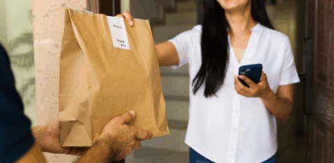 A smiling woman stands at her doorway, accepting a paper bag of food delivery from a courier while holding her phone.