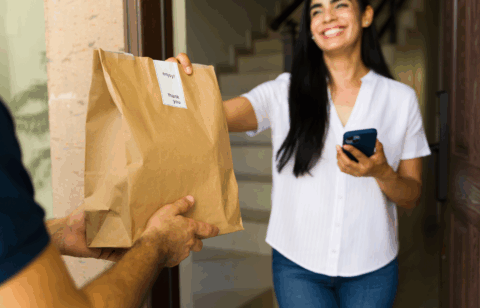 A smiling woman stands at her doorway, accepting a paper bag of food delivery from a courier while holding her phone.