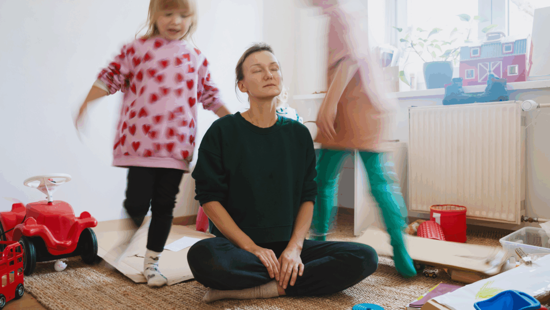 Mother sitting on the floor with eyes closed, trying to relax while children play around her in a messy room.