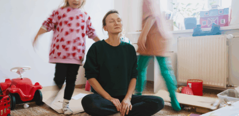 Mother sitting on the floor with eyes closed, trying to relax while children play around her in a messy room.