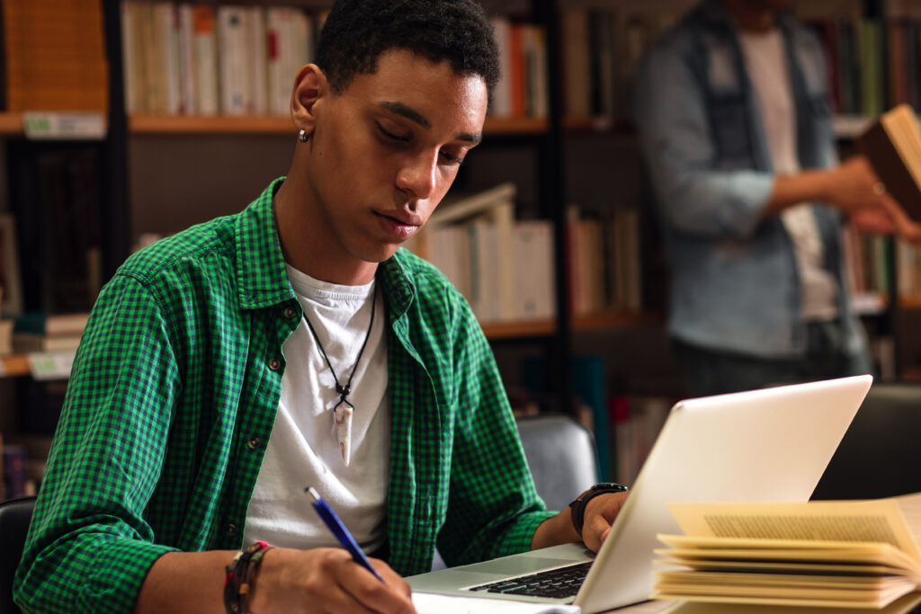 Student studying with a laptop and notebook in a library.