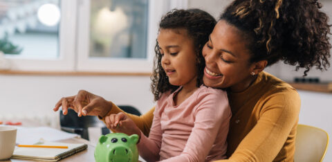 A mother and daughter smile while putting coins into a green piggy bank at the kitchen table.