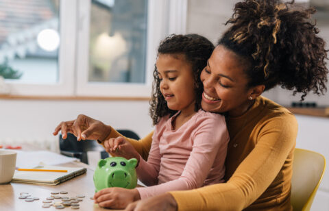 A mother and daughter smile while putting coins into a green piggy bank at the kitchen table.